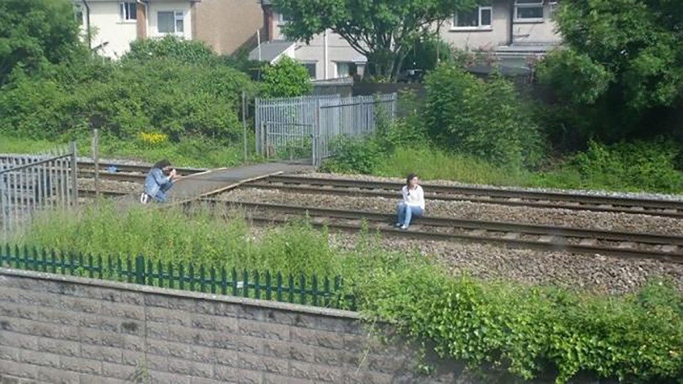  Death trap...Young girl appears oblivious of the dangers of sitting on a track while her friend takes pics on her phone