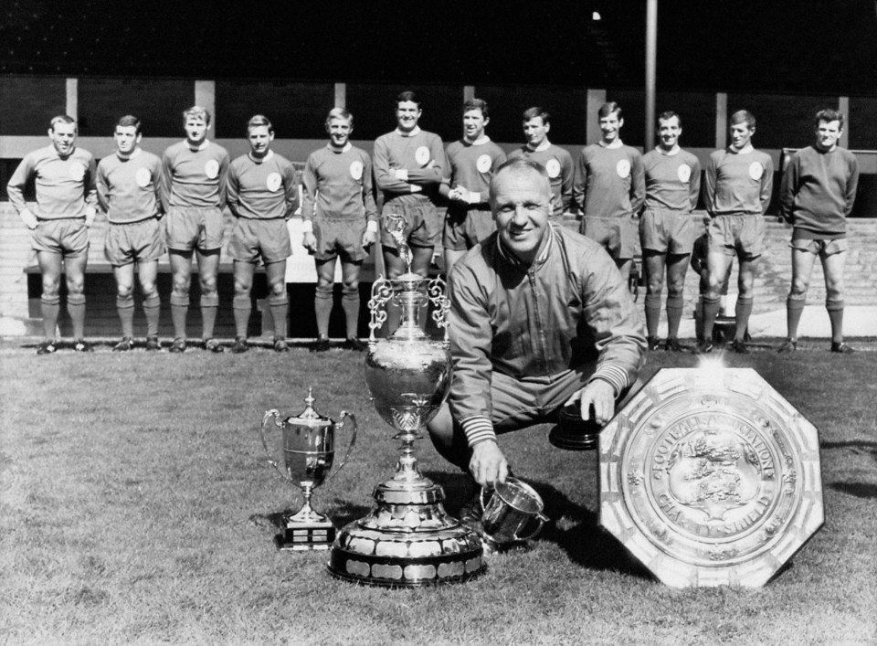 Liverpool manager Bill Shankly crouches by the trophies that his team won the previous season, including the League Championship trophy and the FA Charity Shield, as his players line up in the background: (l-r) Ian St John, Ian Callaghan, Roger Hunt, Gordon Milne, Peter Thompson, Ron Yeats, Chris Lawler, Tommy Smith, Geoff Strong, Gerry Byrne, Willie Stevenson, Tommy Lawrence