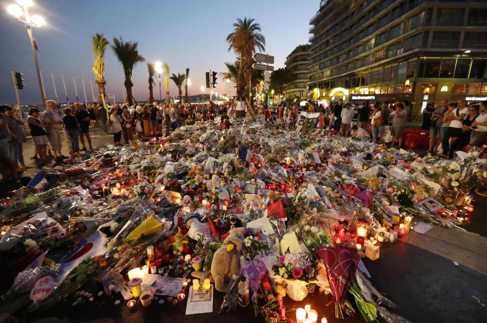 TOPSHOT - People stand in front of flowers, candles and messages laid at a makeshift memorial in Nice on July 18, 2016, in tribute to the victims of the deadly attack on the Promenade des Anglais seafront which killed 84 people. France was set to hold a minute's silence on July 18, 2016 to honour the 84 victims of Mohamed Lahouaiej-Bouhlel, a 31-year-old Tunisian who drove a truck into a crowd watching a fireworks display on Bastille Day, but a period of national mourning was overshadowed by bickering politicians. Church bells will toll across the country, and the country will fall silent at midday, a now grimly familiar ritual after the third major terror attack in 18 months on French soil. / AFP PHOTO / Valery HACHEVALERY HACHE/AFP/Getty Images