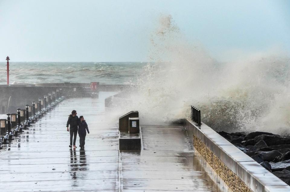  A thrill seeking couple watching a wave crashing over the sea defences of the Jurassic Pier during a morning of rough seas and gale force winds at Dorset's West Bay