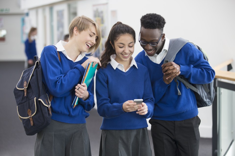 Three smiling students wearing blue school uniforms with mobile phone in school corridor