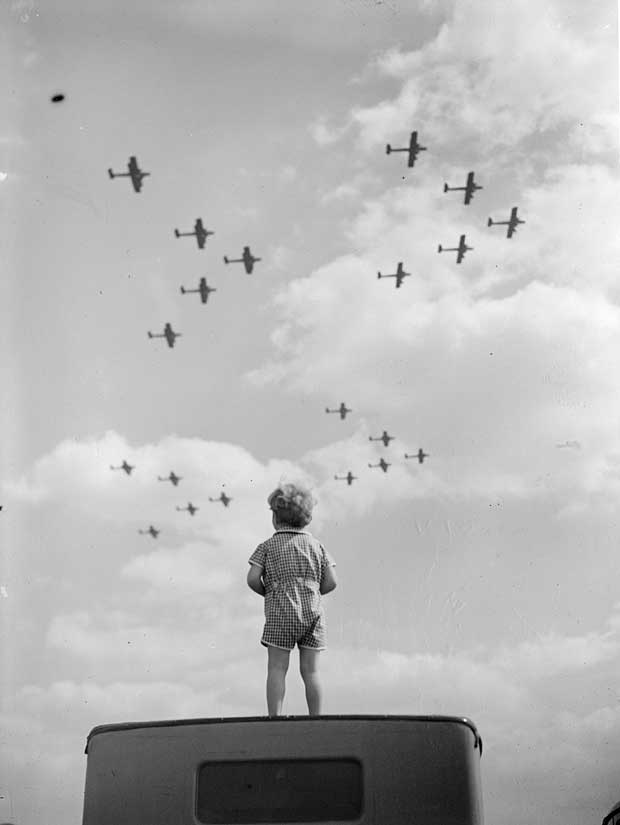  A small boy stands on a car roof to watch RAF warplanes flying in formation.