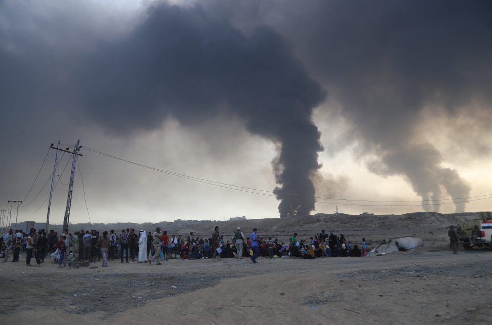 Refugees huddle together while the smoke blows into the sky behind them