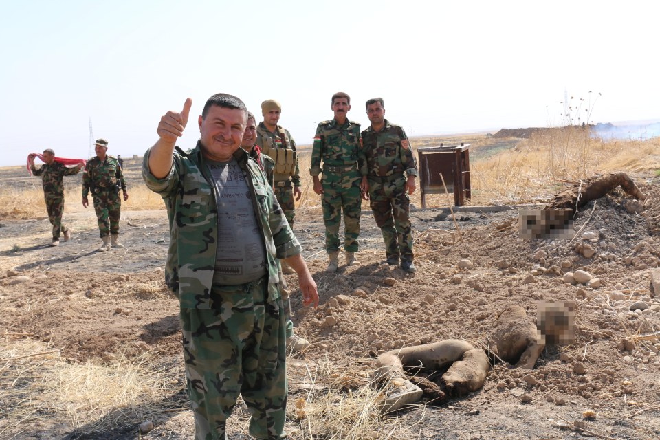 A smiling Kurdish fighter gives a thumbs up next to the body of his enemy