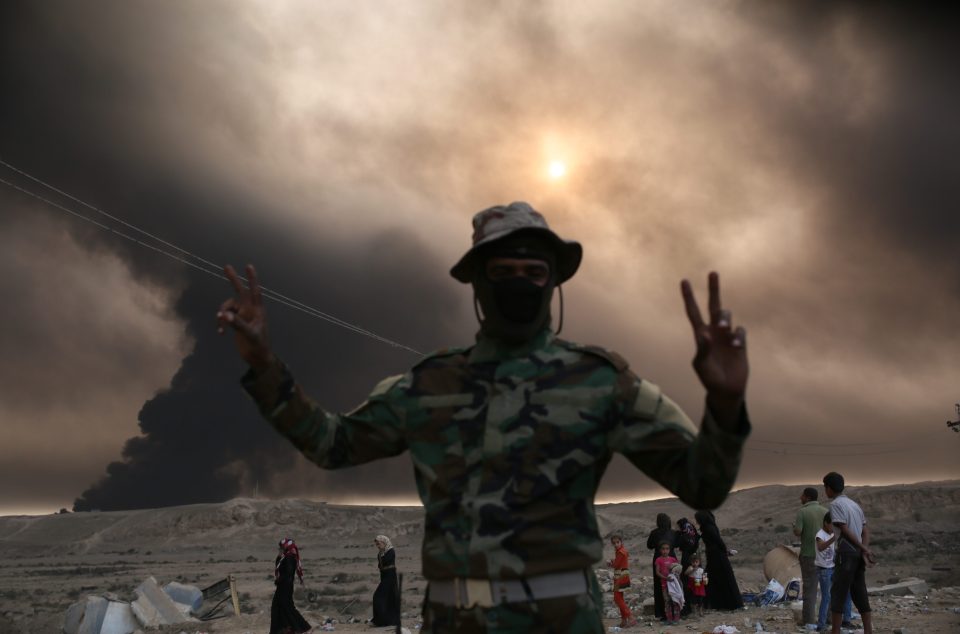  An Iraqi soldier gives the peace sign while standing in front of plumes of toxic smoke