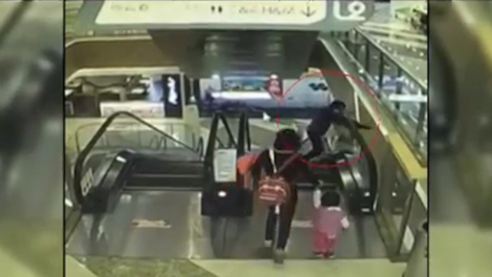  The grandma looks down in horror after the child slips from her arms off the side of escalator