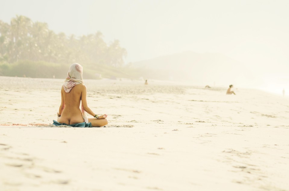 A naked woman is photographed meditating on the beach as the sun begins to set