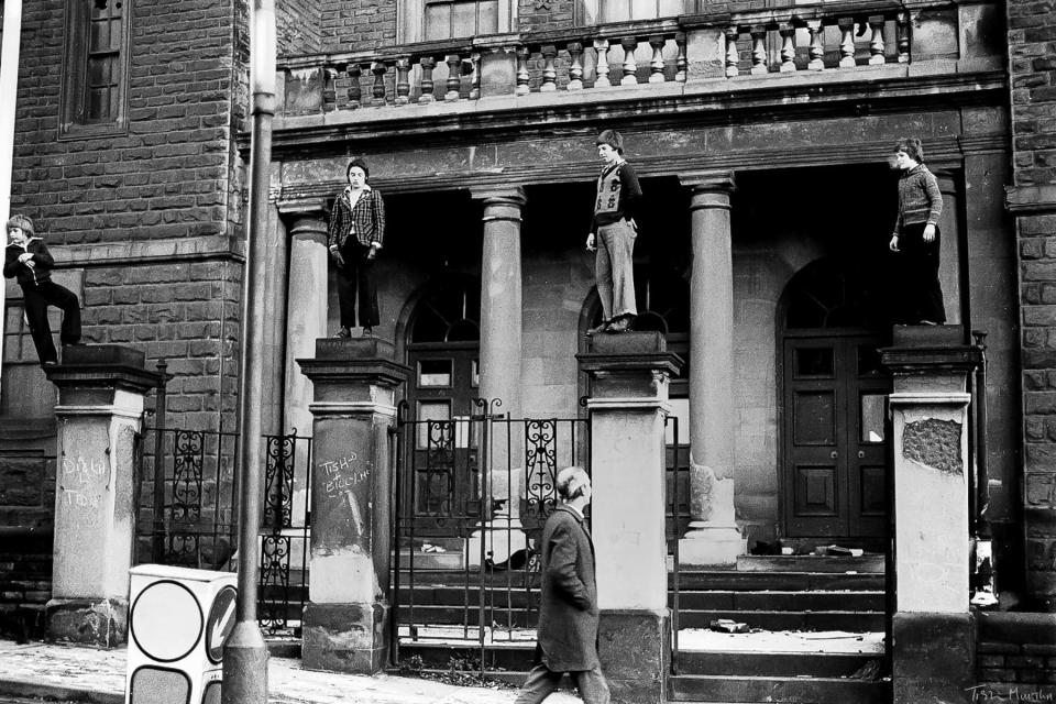  A man gazes at four boys who are balanced on old pillars outside a crumbling building