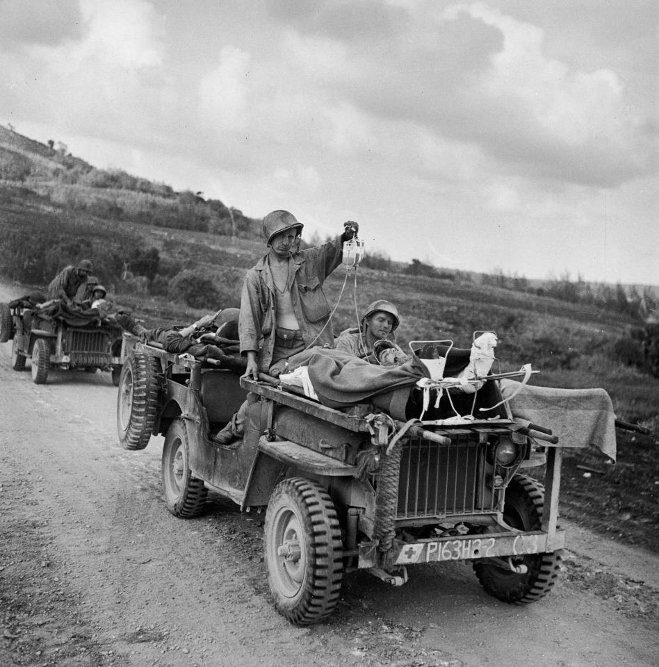  1944: US soldiers drive wounded away from the front line in Saipan, with one trooper holding up IV bags attached to two injured men