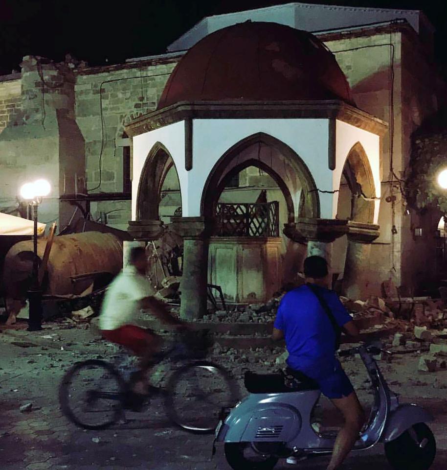  People on bikes and mopeds pass by damaged buildings on Kos in the early hours of this morning