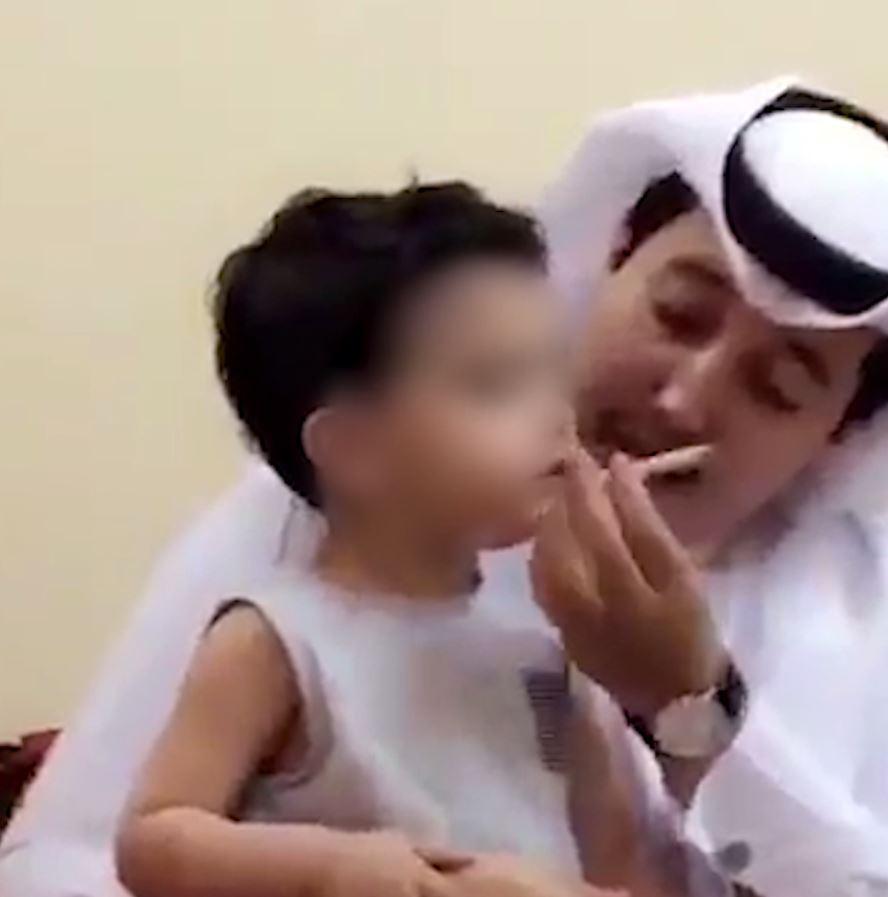  The man places the cigarette in the child's mouth as he sits on his knee