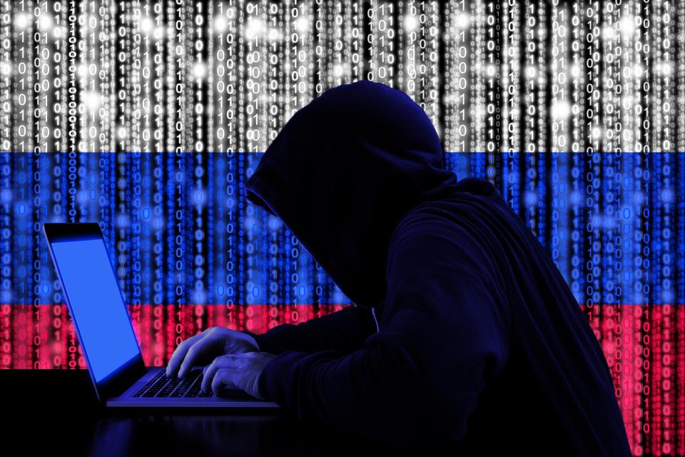 Hooded figure using laptop with Russian flag binary code background.