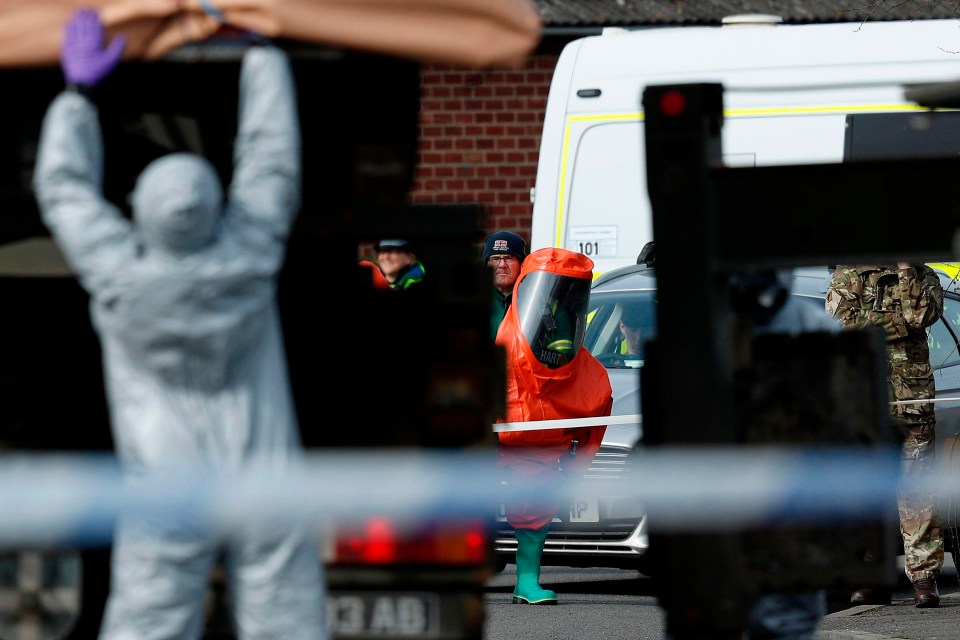 A member of the British ambulance service's Hazardous Area Respose Team wears a red biohazard encapsulated suit as he watches military personnel wearing protective coveralls work to remove the vehicle 