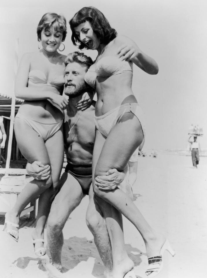  American actor Kirk Douglas, wearing a swimsuit, lifts two women wearing bikinis on a beach during the Venice Film Festival in 1953