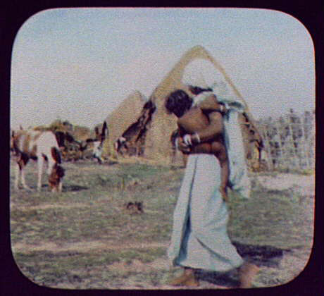  In a village of thatched houses, a women holds her baby and watches a pony