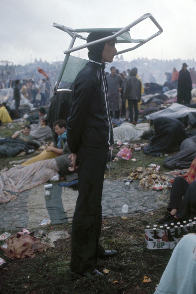  A concertgoer in a raincoat and hood stands with his hands clasped behind his back and a folding deck chair balanced on his head at the three-day event