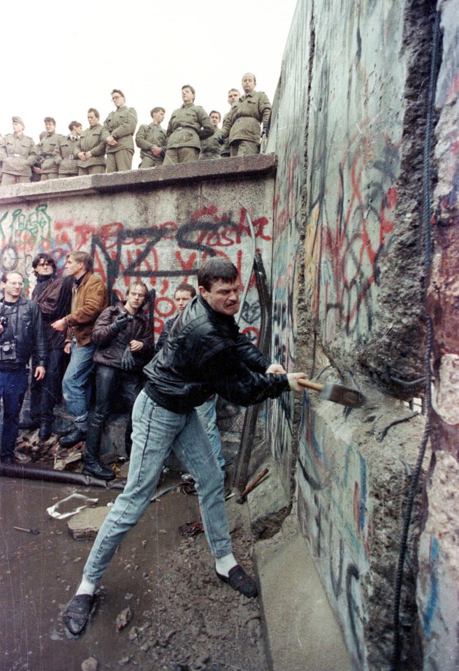  A man pounds away the Berlin Wall as East Berlin border guards look on from above the Brandeburg Gate on November 11, 1989