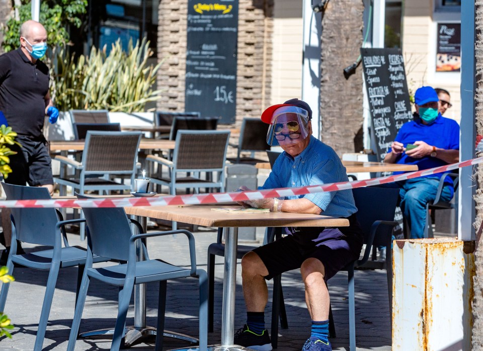  A man with a face visor and mask sits on a bar terrace