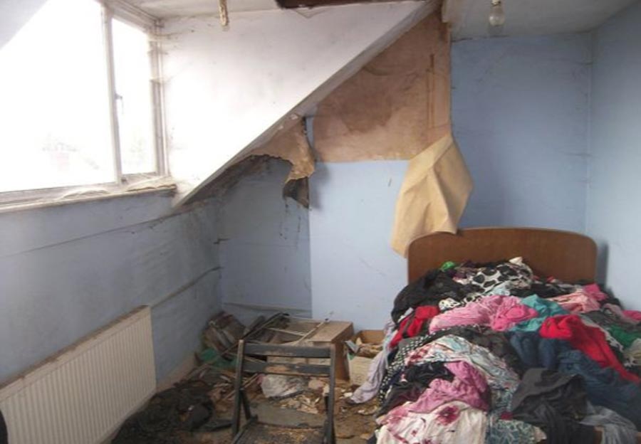 The floor of the second bedroom is covered with debris, with clothes abandoned on the single bed and wallpaper peeling off 