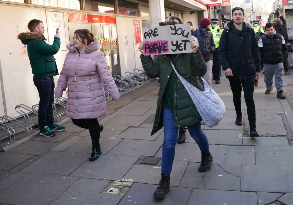 A woman walks through the street with a sign reading 'are you awake yet'