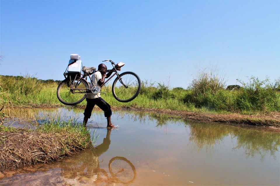 A community relay crosses a water stream with his bike to vaccinate children in the health zone of Manono, Tanganyika Province, in Democratic Republic of the Congo in 2018