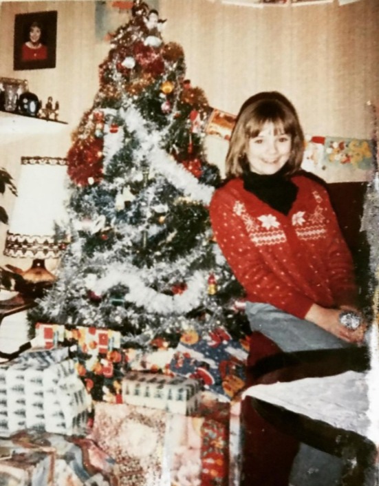 a girl in a red sweater sits in front of a christmas tree