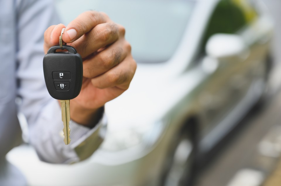 A hand holding a car key with a blurred car in the background.