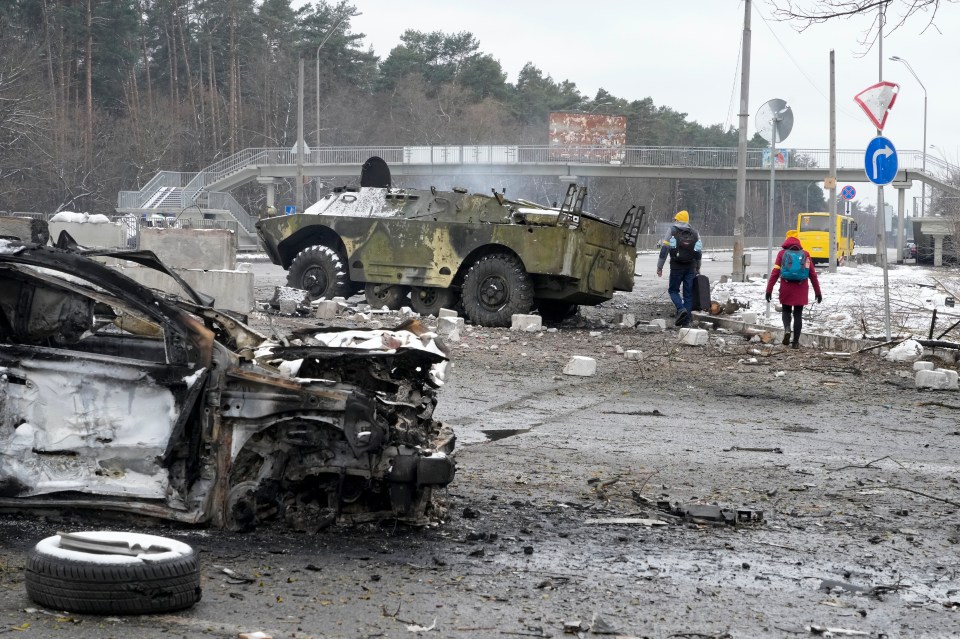 People walk by a damaged vehicle and an armoured car at a checkpoint in Brovary, outside Kyiv