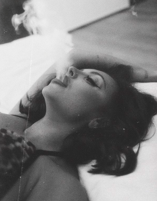 She puffs smoke while stretched out on a bed in another