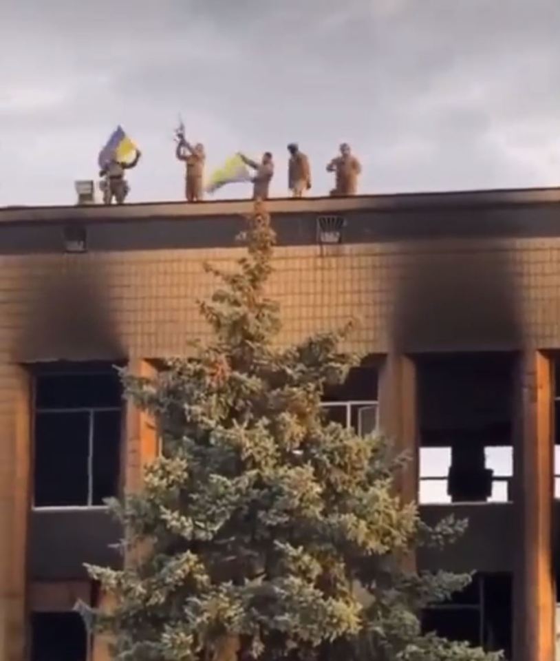 Five Ukrainian servicemembers hold flags and fire guns in air possibly from top of City Council building in Izium