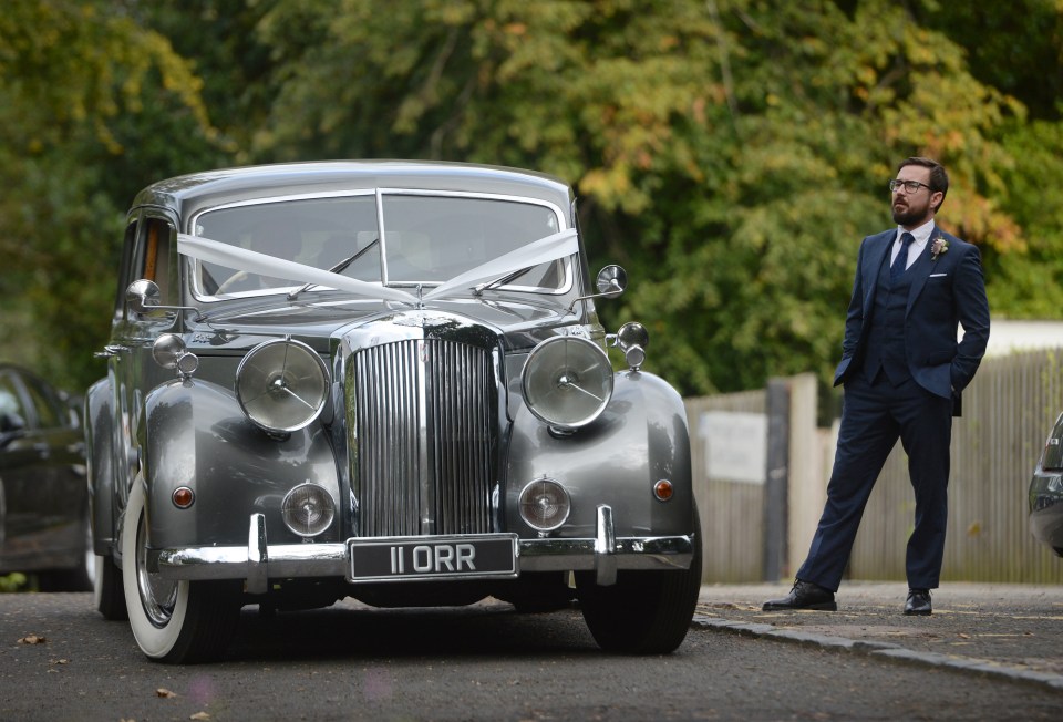 He sports a navy three-piece as he stands alongside a wedding car