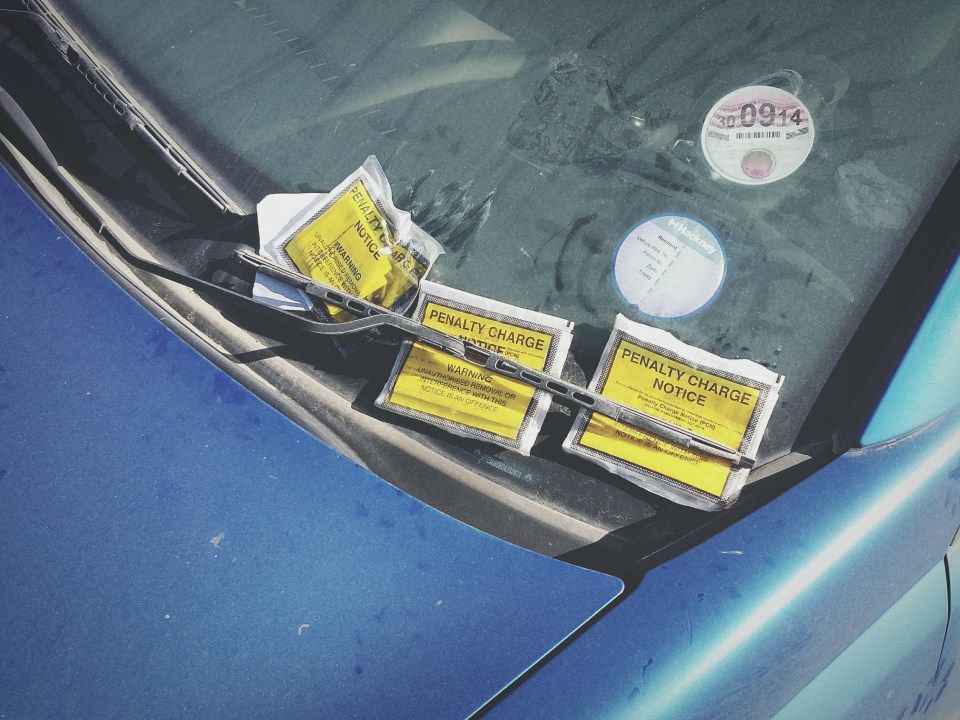 Close up of three parking tickets on the windscreen of a dirty blue car.