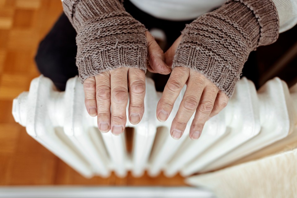 A man hands in wool gloves warm near the heater. Old men’s hands in knitted gloves on heating radiator at home during the day. Person heating their hands at home over a domestic radiator in winter.