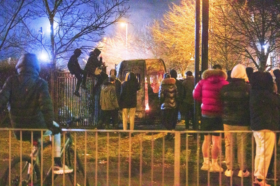 People gather around a police riot van that has been set alight