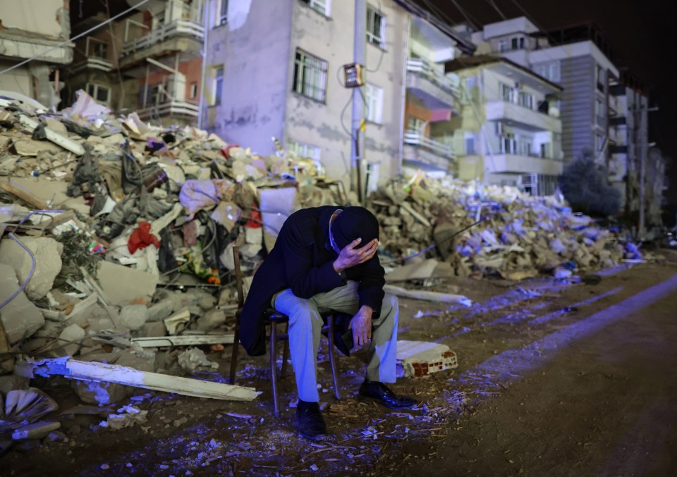 A man holds his head in his hands surrounded by the remains of a destroyed building