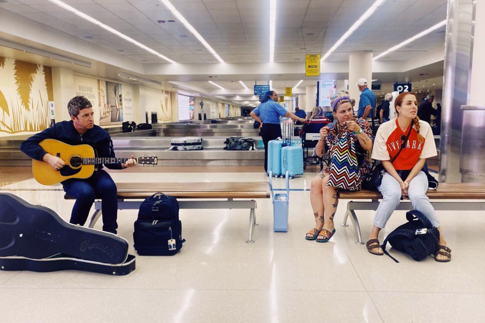 Noel Gallagher strums his guitar at an airport’s baggage reclaim — but two women appear less than impressed