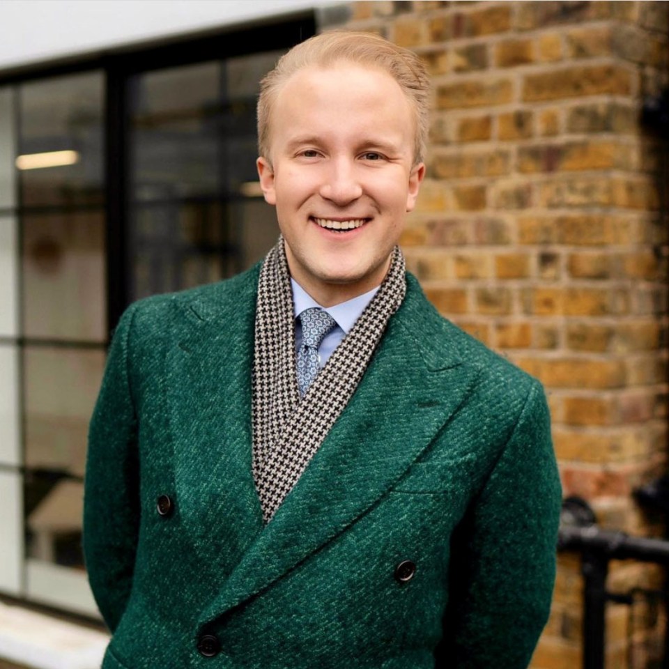 a man in a green coat and scarf smiles in front of a brick wall