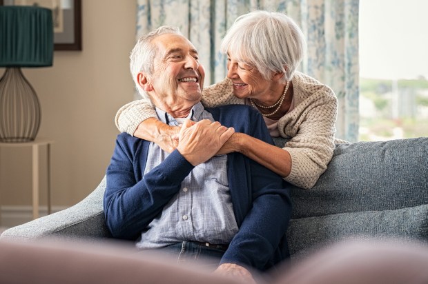 Happy senior woman embracing her husband at home while laughing together. Smiling wife hugging old man sitting on couch from behind. Joyful retired couple having fun at home while looking at each other with copy space. Love and unity concept.