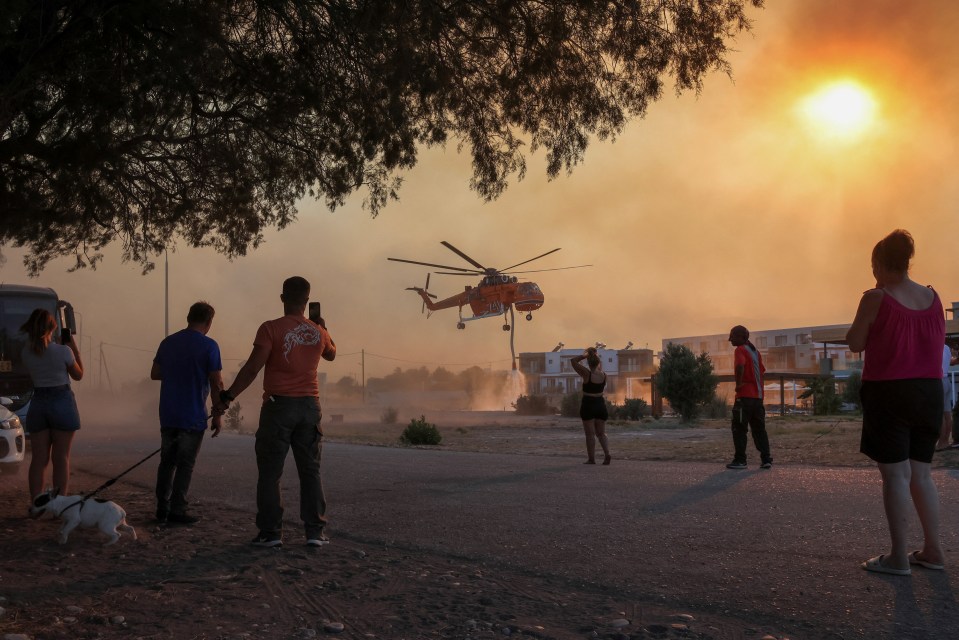 People look at a firefighting helicopter filling water from a pool, as a wildfire burns in the village of Gennadi