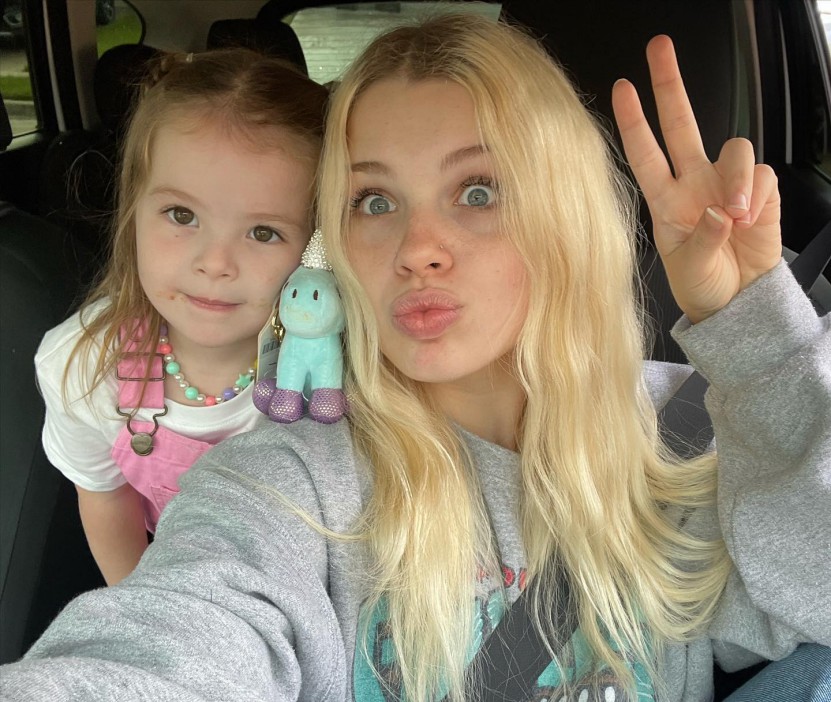 Young woman and toddler girl in a car; the woman is making a peace sign.