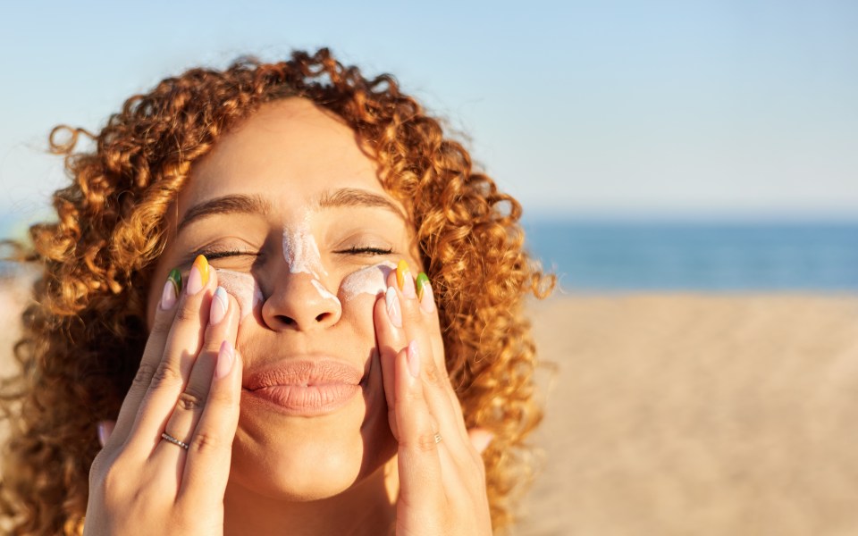 Young latin woman applying sunscreen to her face on the beach in a summer sunset. Cheerful gesture with her eyes closed.