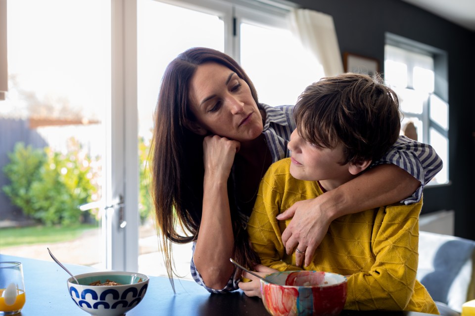 A waist-up shot of a mother reaching her arm around her son next to the kitchen counter looking unhappy with her son. They are wearing casual clothing.