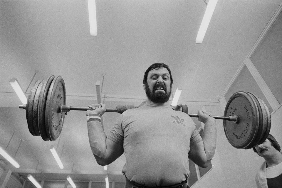 Black and white photo of strongman Geoff Capes straining to lift a barbell.