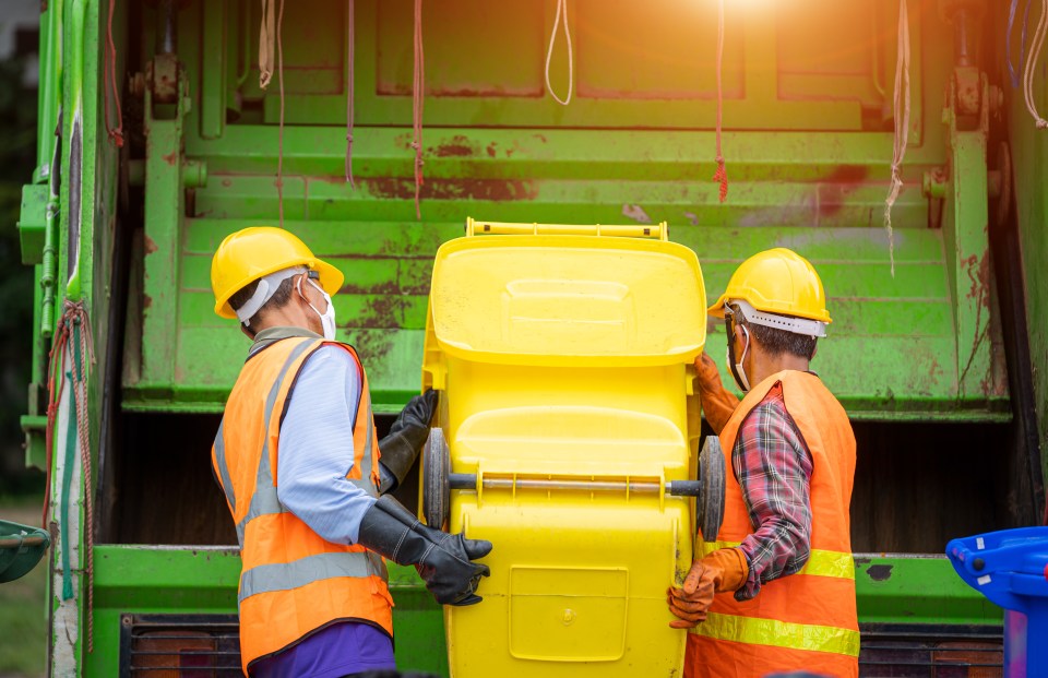 two men wearing hard hats and orange vests are loading a yellow trash can into a green garbage truck