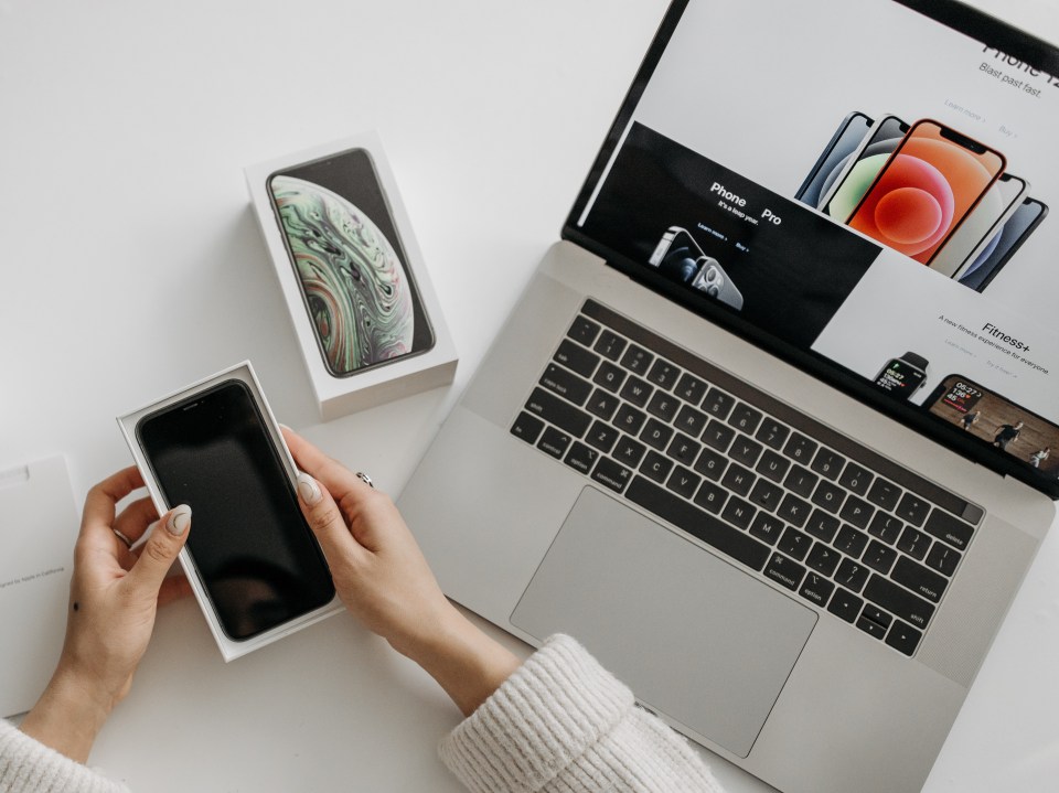 Person holding a new iPhone in its box, next to a laptop showing an Apple website.