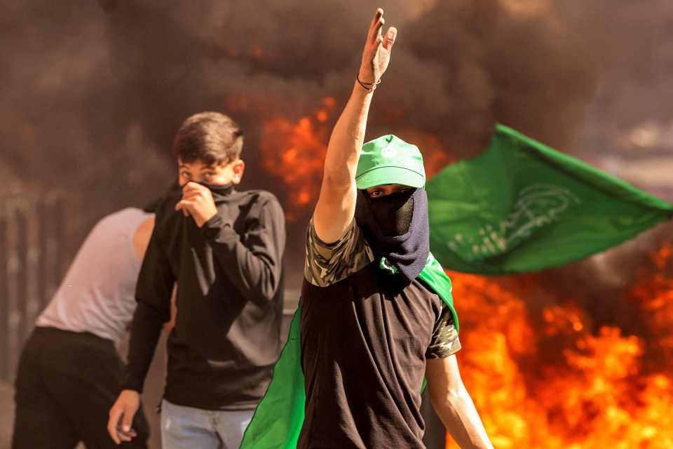 A masked Palestinian protester gestures while walking near flaming tires in the West Bank