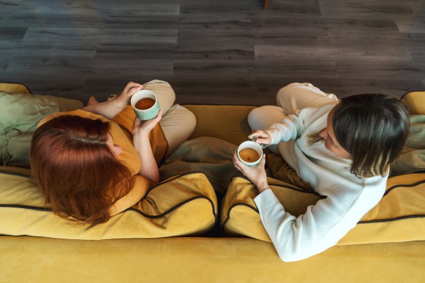 two women sit on a yellow couch drinking coffee