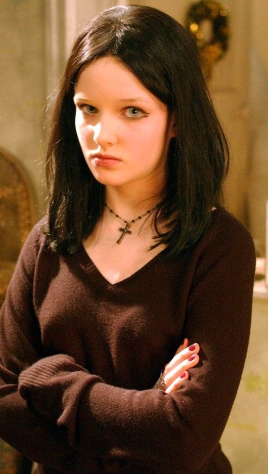 a girl wearing a cross necklace and a brown sweater has her arms crossed