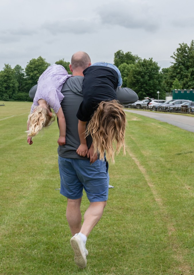 A man carrying two young girls on his shoulders walks across a grassy field.