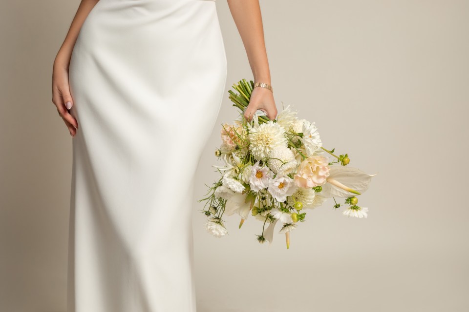 Unrecognizable cropped woman in white elegant silk dress with white bouquet of natural flowers in hand with accessories on light studio background.